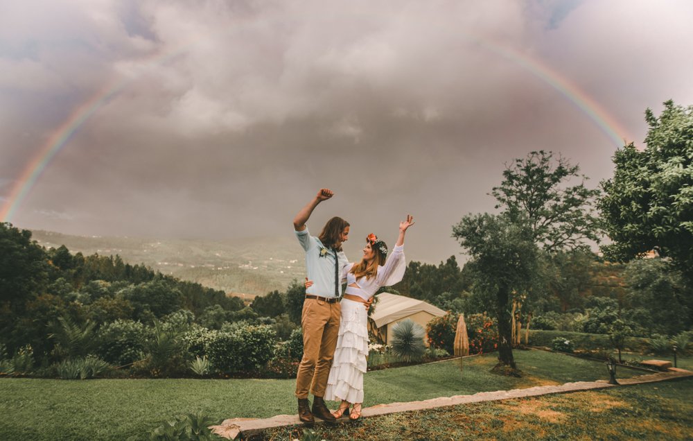 Bride in boho chic dress with flower crown standing with groom under a rainbow on their rainy wedding day
